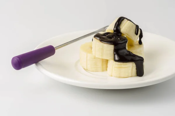 Doğranmış muz çikolata sır fondü çatal ile bir tabak içinde bulunmaktadır