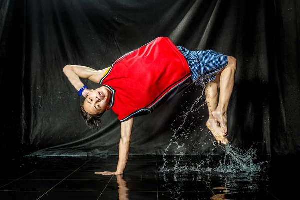 cool and happy korean young guy break dancer in style of bboying doing complex tricks on floor with water splashes and liquid drops effects in aqua zone photo studio with black isolated background