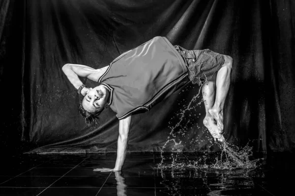 cool and happy korean young guy break dancer in style of bboying doing complex tricks on floor with water splashes and liquid drops effects in aqua zone photo studio with black isolated background