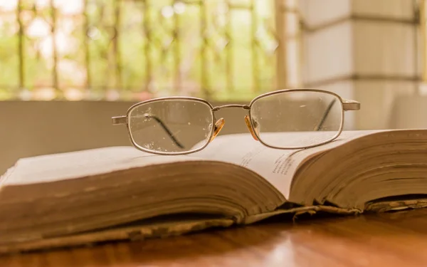 Un libro aperto sul tavolo e uno specs o occhiali da vista sul tavolo di legno, in una calda mattina di sole, vista laterale da vicino. Educazione o concetto di vacanza. Fondo isolato a fuoco selettivo, profondità di campo poco profonda — Foto Stock