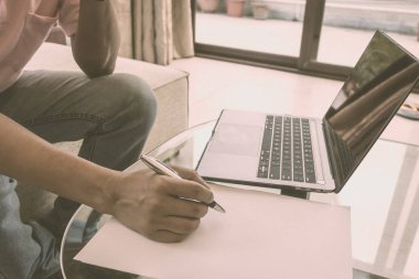 Anonim İşçi bir kağıt üzerine bir iş projesi yazıyor. Ofis masasında. Laptop masanın üzerine yerleştirildi. Yan görüş. Yakın plan. Klasik tarz etkisi. Metin için boşluk kopyala.