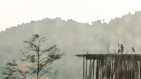 Неузнаваемая молодая женщина на дальнем расстоянии, стоящая на крыше старого строящегося здания, окруженная живописной тенью величественной Гималайской горной долины и соснового леса
.