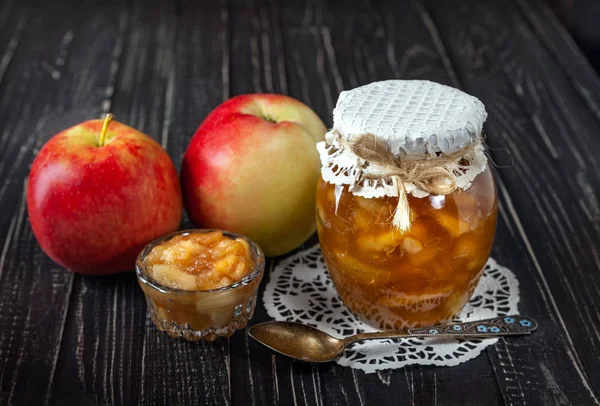 Small dish with jam and jar with apple jam with apples and a spoon on a black wooden table