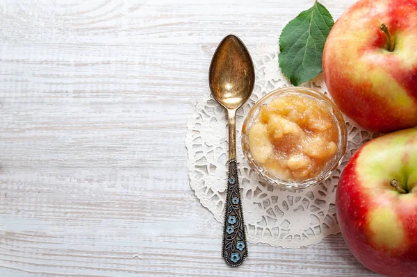 Small dish with apple jam with whole apples and a spoon on a white wooden table, selective focus