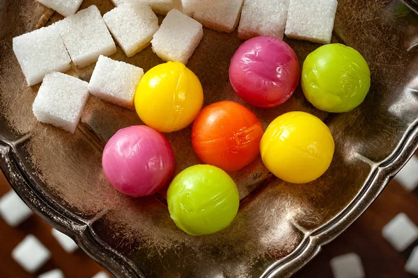 Candy balls and pieces of sugar. Colored dragees hard fruit candy and sugar stacked on a silver saucer, wooden background. The concept of sweet life.