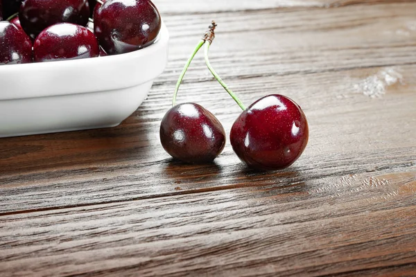 白いプレートに赤い新鮮な熟したジューシーな甘いサクランボと木製の背景に2つの果物。水滴と濡れる甘いチェリー. — ストック写真