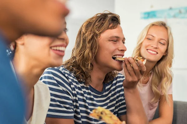 Portrait of a young man biting a slice of pizza at home with friends. Happy smiling beautiful women and men having pizza party. Group of girls and guys sitting on sofa and having fun time together.