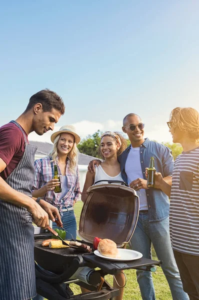 Group of multiethnic young friends enjoying barbecue in the backyard. Happy men and beautiful women talking and grilling meat. Smiling guys and girls having a good time at outdoor party during sunset and drinking beers.
