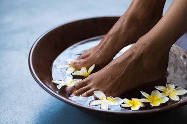 Woman soaking feet in bowl of water with floating frangipani flowers at spa. Closeup of a female feet at wellness center on pedicure procedure. Woman feet in spa wooden bowl with exotic white flowers. clipart