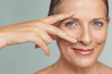 Zafer işareti eye yakınındaki gri arka plan üzerinde gösterilen mükemmel cilt ile gülümseyen kadın kıdemli portresi. Olgun kadın anti-aging kırışıklık tedavi sonrası başarılı sonuçlar closeup yüzü. Güzellik olgun cilt bakım kavramı.