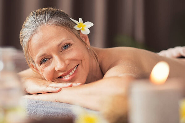 Beautiful mature woman lying on front on a massage table and looking at camera. Portrait of senior woman for a beauty treatment in a spa center. Skin care and body massage concept.