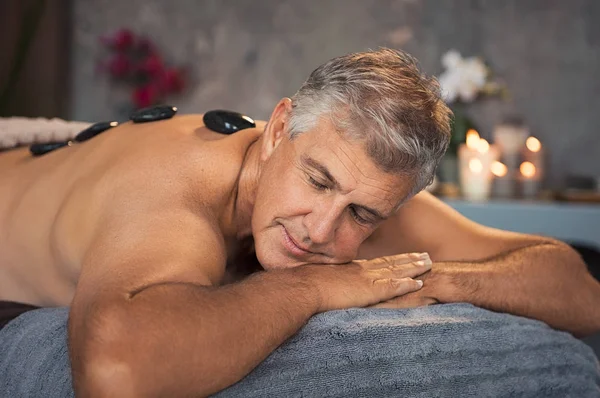 成熟した男がホットな石のマッサージ中にベッドでリラックス 温石療法中に休んで目を閉じて年配の男性の肖像画 バック グラウンドでの蝋燭とスパで背中に黒い溶岩石でハンサムな男 — ストック写真