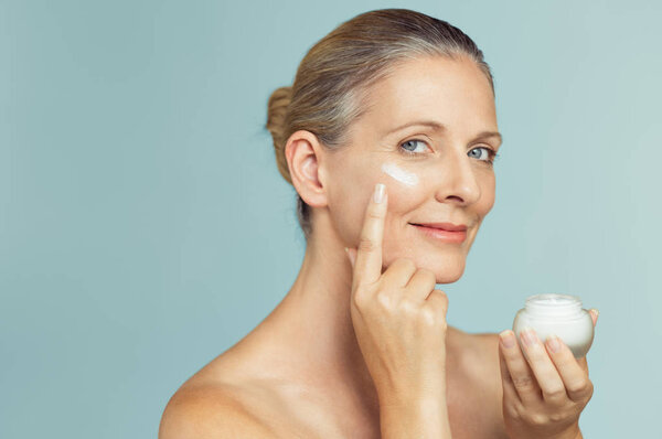 Beautiful mature woman holding jar of skin cream for face and body isolated on grey background. Happy senior woman applying anti-aging moisturizer and looking at camera. Beauty and anti aging treatment.