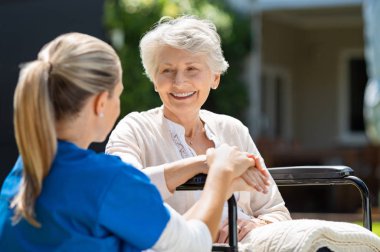 Smiling senior patient sitting on wheelchair with nurse supporting her. Doctor looking at elderly patient on a wheelchair in the garden. Nurse holding hand of mature woman outside pension home. clipart