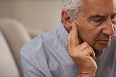 Side view of senior man with symptom of hearing loss. Mature man sitting on couch with fingers near ear suffering pain. clipart