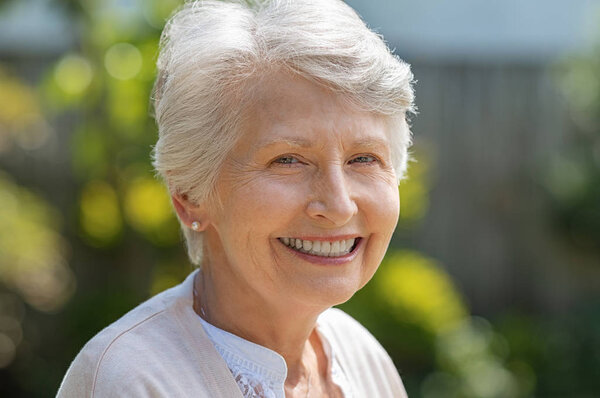 Portrait of serene old woman in garden looking at camera. Closeup portrait of happy older beautiful woman standing outside in summer and smiling. Happy retired senior woman relaxing outdoor. 