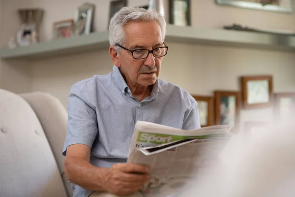坐在沙发上看报纸的老人画像 退休男子戴眼镜 而在家里阅读新闻 资深祖父阅读坏消息 — 图库照片