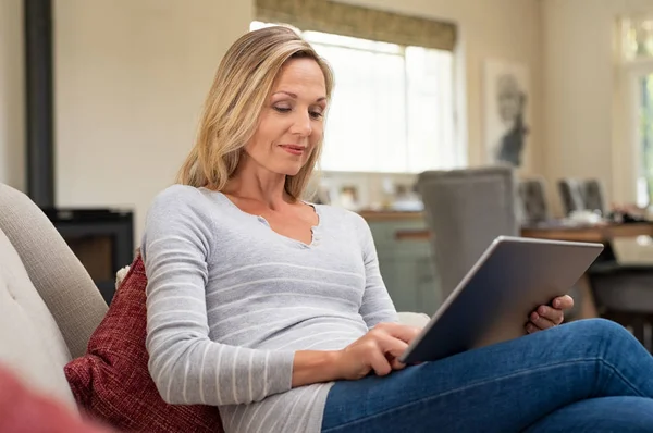 漂亮的成熟女人坐在沙发上 用数码片 微笑的女士浏览互联网在平板电脑在家 中年妇女在家里拿着电脑 — 图库照片