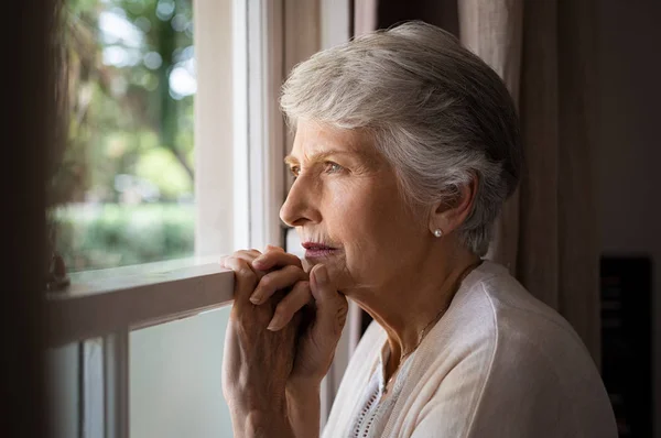 郁闷的老妇人在家里感到难过 老妇人悲伤地看着窗外 孤独的忧郁的女士独自站着 透过窗户看着 — 图库照片