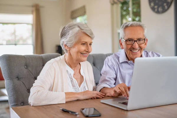 幸福微笑的退休夫妇在家里使用笔记本电脑 开朗的老人和老太太坐在桌边使用电脑 微笑的领取养老金的女性笔记本在家里显示 — 图库照片