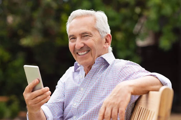 高级男子使用智能手机 而坐在长凳上公园 英俊的老人的肖像使用智能手机户外 快乐微笑的祖父拿着手机看着摄像机 — 图库照片