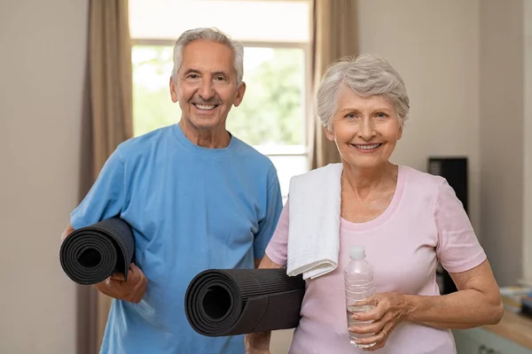 Active healthy senior couple holding yoga mats while going together to training. Happy fitness elderly man and woman looking at camera. Portrait of old couple exercising together at home.
