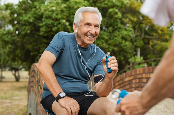 イヤホンと手のグリップを使用して運動の音楽を聴いて幸せな年配の男性 笑みを浮かべて老人が公園で彼の妻を見ながら運動を行います 老人の手の筋肉を強化するトレーニング — ストック写真
