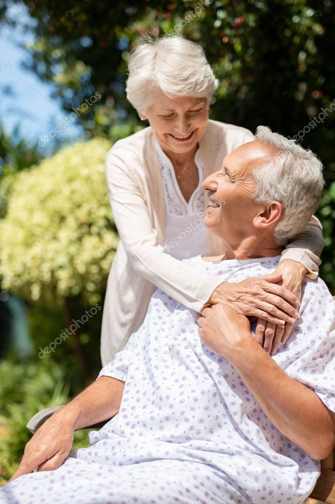 Senior woman hugging man in medical cloth in the hospital garden. Loving wife embracing old hospitalized husband sitting on bench at outdoor lawn. Caring wife supporting husband in illness in a private medical clinic.