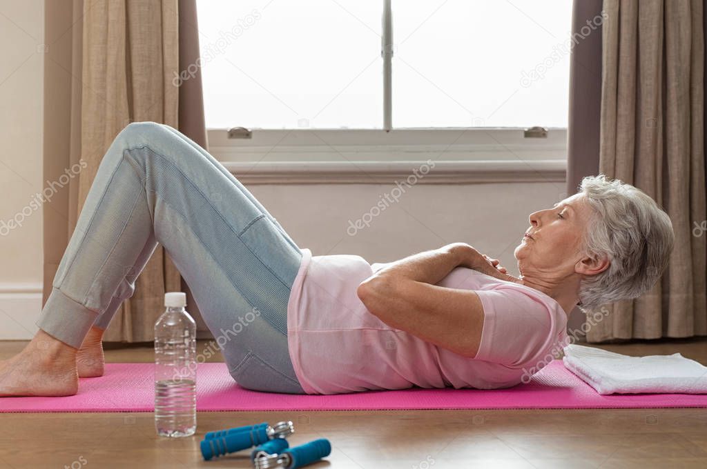 Senior woman doing sit ups on yoga mat at home. Fit healthy old woman doing abdomen crunches at home. Elderly woman keeping fit by exercising for staying healthy.