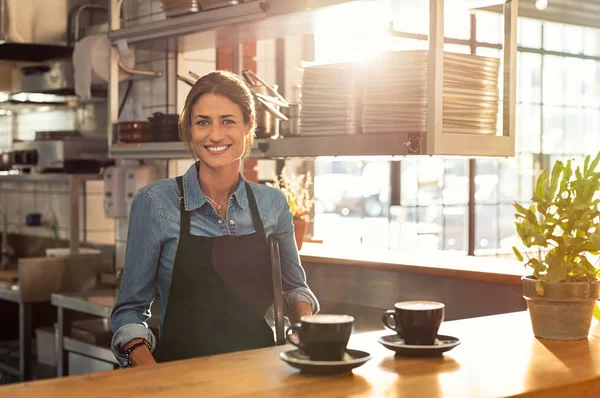 微笑的女服务员穿着黑色的围裙站在柜台后 在自助餐厅和看着相机 在自助餐厅为顾客提供两杯热咖啡的成熟女人 小企业与企业家理念 — 图库照片