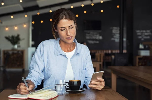 成熟した女性のテーブルでカフェに座っているとスマート フォンでメッセージを入力します 中年ブロガー電話で組織化アプリケーションを使用してノートを作るします カフェのインテリアでリモートでの作業中 スマート フォンから情報を読み取って実業家 — ストック写真
