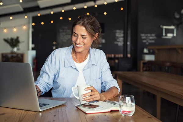 坐在自助餐厅的微笑的女人拿着咖啡杯 在笔记本电脑上工作 女商人在笔记本电脑上检查电子邮件 美丽的中年妇女微笑和使用笔记本电脑在咖啡馆 而喝一杯茶 — 图库照片
