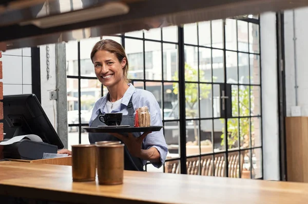 Smiling waitress serving cup of coffee in restaurant. Happy woman holding tray with cappuccino at the coffee shop and preparing bill while looking at camera. Beautiful woman holding tray with coffee making cheque for customer.