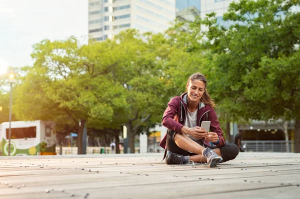 移动智能手机应用程序跟踪进度的健身跑步者 运动员成熟妇女在运动装听音乐用耳机在城市街道 拉丁运动妇女坐在地板上 并使用智能手机与复制空间 — 图库照片