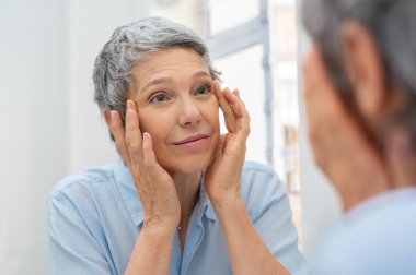 Beautiful senior woman checking her face skin and looking for blemishes. Portrait of mature woman massaging her face while checking wrinkled eyes in the mirror. Wrinkled lady with grey hair checking wrinkles around eyes, aging process. clipart