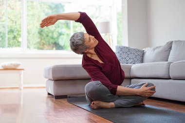 Lotus bulunduğu otururken egzersiz kadın kıdemli. Etkin Olgun kadın evde oturma odasında germe egzersiz yapmak. Bayan Kol germe uygun ve yoga mat üzerinde otururken geri.