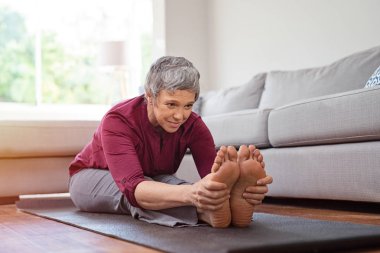 Evde yoga mat üzerinde otururken germe egzersiz yaparak güzel kadın kıdemli. Olgun kadın spor içinde ileri germe egzersiz ayak dokunmak. Yoga ve esneklik egzersiz yaparak sağlıklı aktif Bayan.