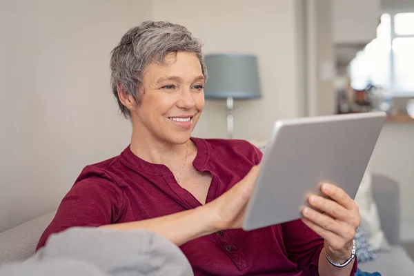 微笑的资深妇女坐在沙发上看着她的数码平板电脑 在家里用数码片放松的成熟快乐女人的肖像 带灰色头发的快乐女士在起居室的笔记本电脑上浏览 — 图库照片