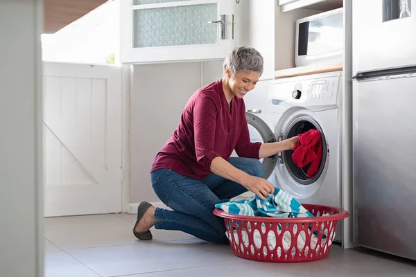快乐的资深妇女在洗衣机里装脏衣服 微笑着成熟的女人坐在地板上 把衣服从洗衣篮装进洗衣机里 — 图库照片