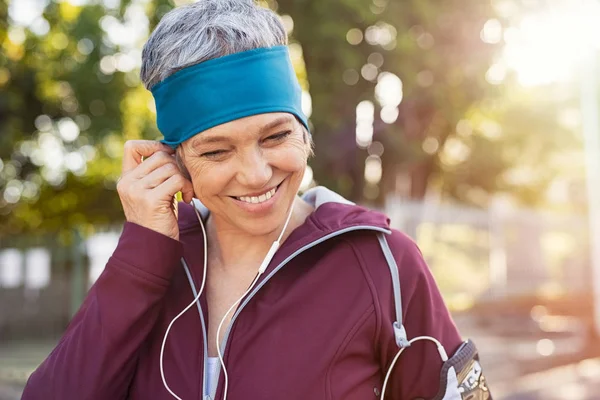 微笑的资深妇女的画像在奔跑以后听音乐 健康成熟的妇女身穿蓝色头巾和运动装调整耳机 而慢跑 退休女子在公园慢跑的特写脸 — 图库照片