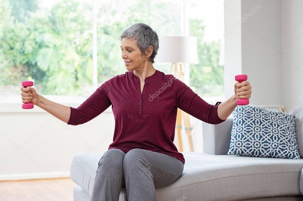 Senior woman lifting weights and working out at home. Mature woman sitting on couch doing hand stretching exercise using light weight dumbbells. Beautiful old lady exercising at home to stay fit.