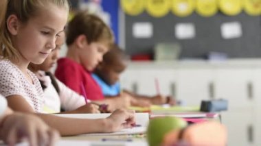 Mutlu güzel kız öğrenci kameraya bakarken classwork yapıyor. Arka planda sınıf ile okul masasında oturan bilgin kız gülümsüyor. Genç oldukça zevk çalışmada İlköğretim Okulu çocuk.