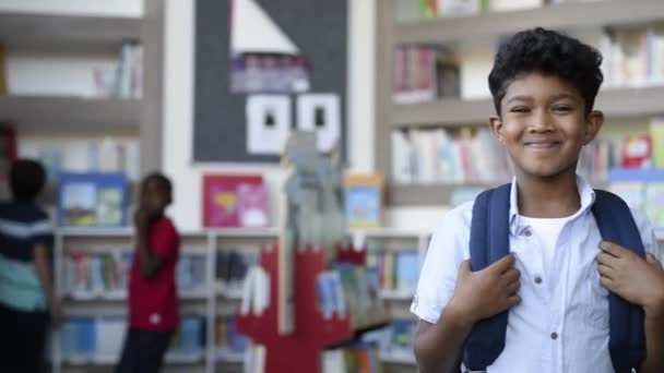 面带微笑的惊慌失措的男孩看着相机的画像 小学生背着背包 站在学校图书馆里 带着图书馆背景的快乐的中东儿童 — 图库视频影像