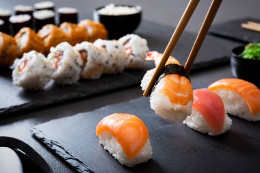 Karides suşi nigiri ahşap yemek çubukları ile siyah bir tabakta. Yemek çubukları ve soya sosu ile uramaki ve hosomaki arka planda ile oluşturulmuş nigiri closeup. Restoranda siyah arduvaz yemekleri Japon yemek yeme.