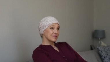 Olgun kadın evde kanepede oturan ve uzağa bakarak gülen pembe başörtüsü kanserli. Orta kadın kanser kemoterapi oturumları kameraya bakarak aldıktan sonra oturup acı. Üzgün kadın saç kaybı bakarak yan etkilere karşı karşıya portresi 