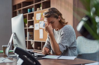 Yorgun işkadını modern ofiste having bir baş ağrısı. Ofis Masası duygu yorgun baş üzerine gözlük ile çalışan olgun yaratıcı kadın. Masaüstü bilgisayarda overworking süre rahat iş kadın duygu göz ağrısı vurguladı.