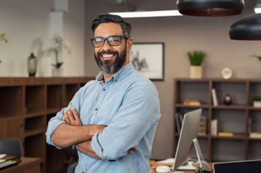 Gözlük takan ve kameraya bakarak mutlu olgun işadamı portresi. Sakal ve gözlük ofiste duygu kendinden emin çok ırklı memnun adam. Yaratıcı bir ofiste gülümseyen başarılı Orta Doğu iş adamı.