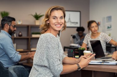 Olgun işkadını iş toplantı sırasında Smartphone kullanarak. Postaları kontrol ve mobil kameraya bakarken yanıtlarken başarılı bir iş kadını. Ofiste akıllı telefon kullanarak gülümseyen casual Bayan portresi.