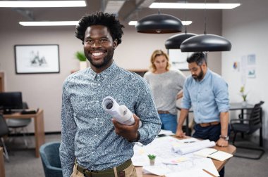 Neşeli Afrikalı mimar planları tasarım rulo tutarak. Mutlu mühendis arka planda çalışma ekibi ile ayakta portresi. Yeni mimari projede çalışan başarılı Afro-Amerikan casual adam.