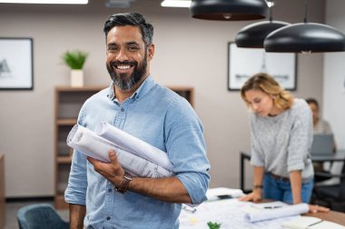 Çok ırklı mimar planları yaratıcı Office ile portresi. Mimari projeler rulo kameraya bakarken tutan olgun Orta Doğu müteahhit. Mutlu latin adam backgr içinde çalışma mühendisleri ile Toplantı salonunun içinde casual ayakta